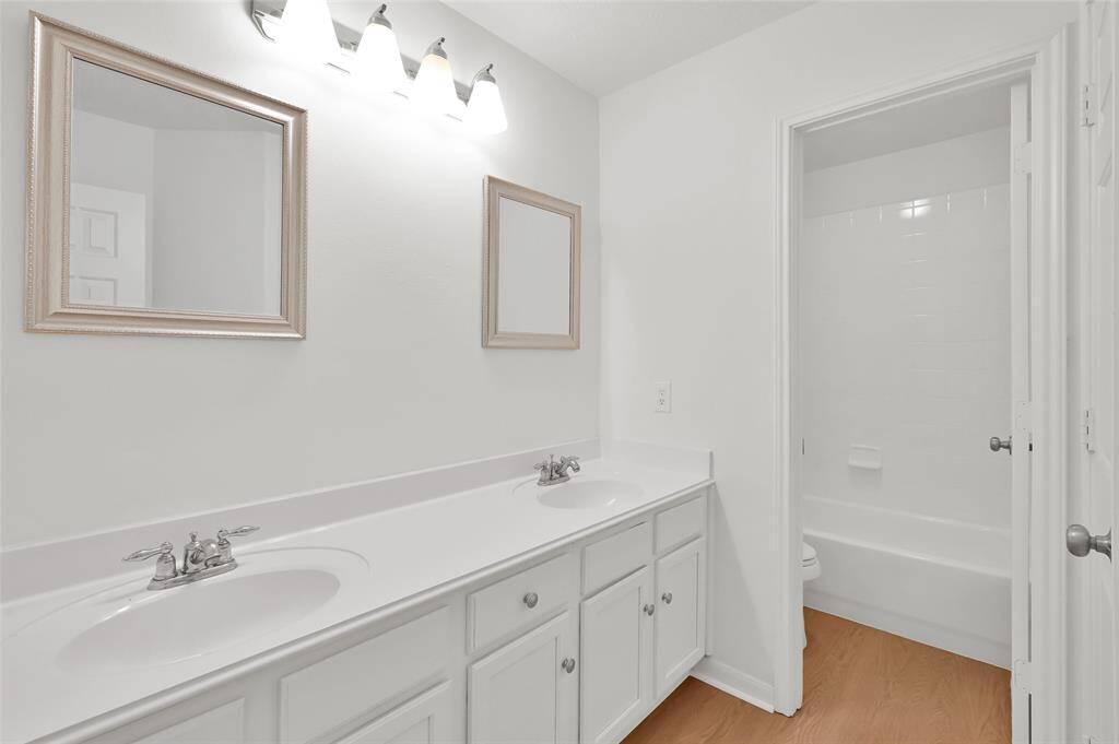 This is a bright, modern bathroom featuring a double vanity with two sinks, framed mirrors, and ample cabinet storage. It includes a bathtub with a shower in a separate alcove, and has a clean, white color scheme with wood-like flooring.