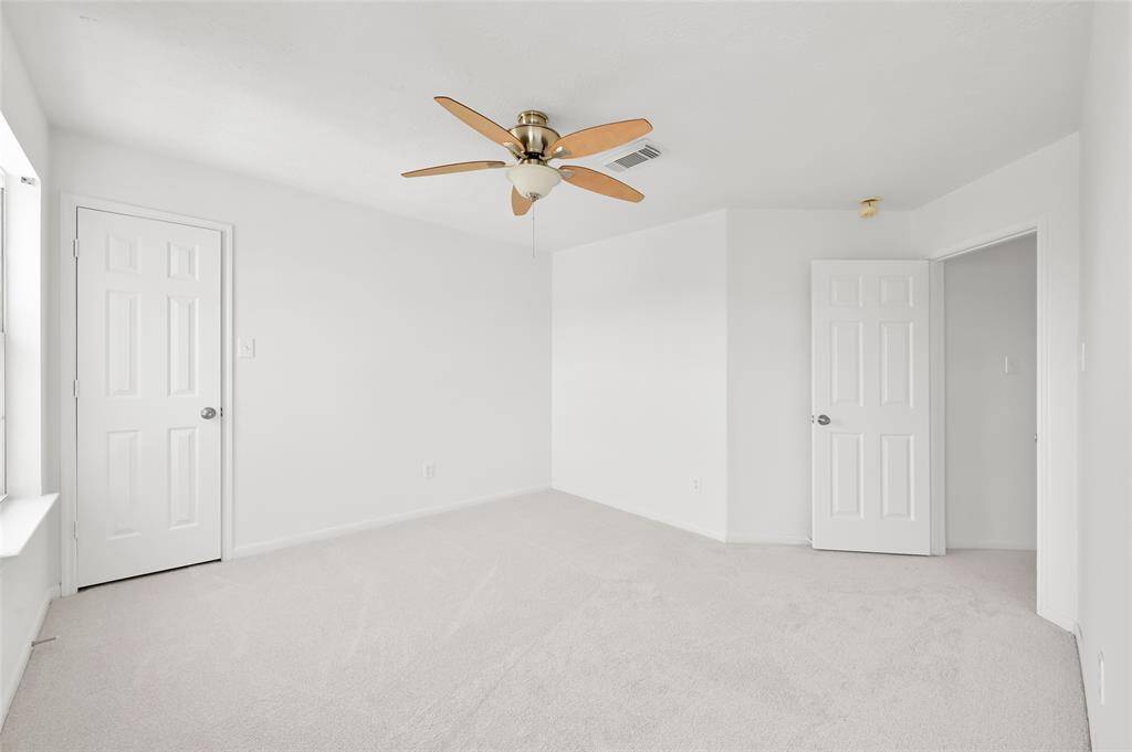 Bright, spacious bedroom with neutral carpeting, white walls, and a ceiling fan. It features two doors, one leading to a closet and leading to the hall, and ample natural light from a large window.