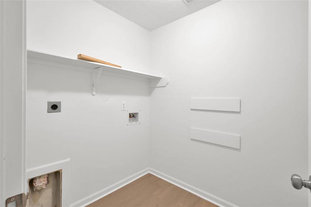 This photo shows a laundry room with white walls, a single shelf for storage, and hookups for a washer and dryer. The room has a clean, minimalistic design with wood-like flooring.