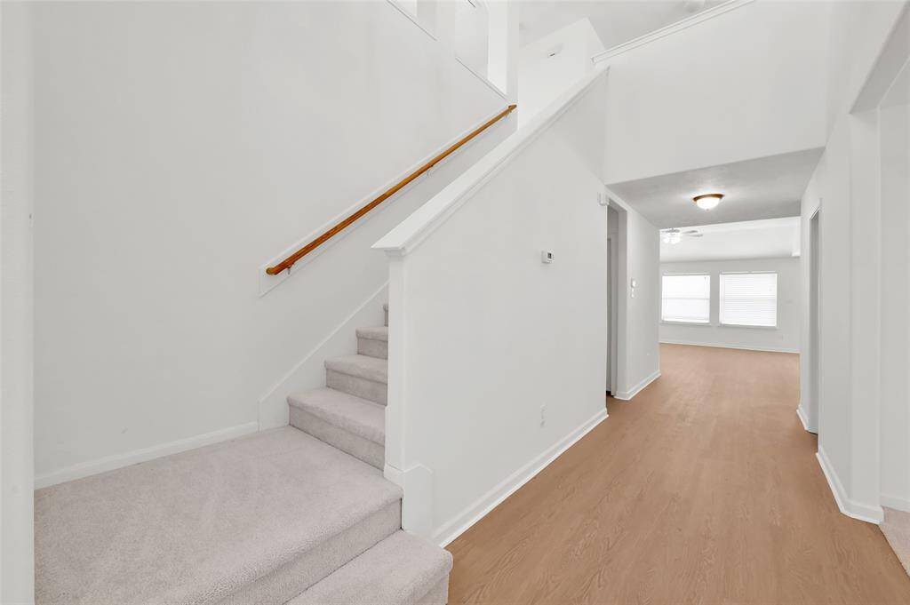 The photo shows a bright, open hallway with light-colored walls and flooring. A carpeted staircase with a wooden handrail leads to the upper floor. The hallway opens into a room with large windows, providing ample natural light.