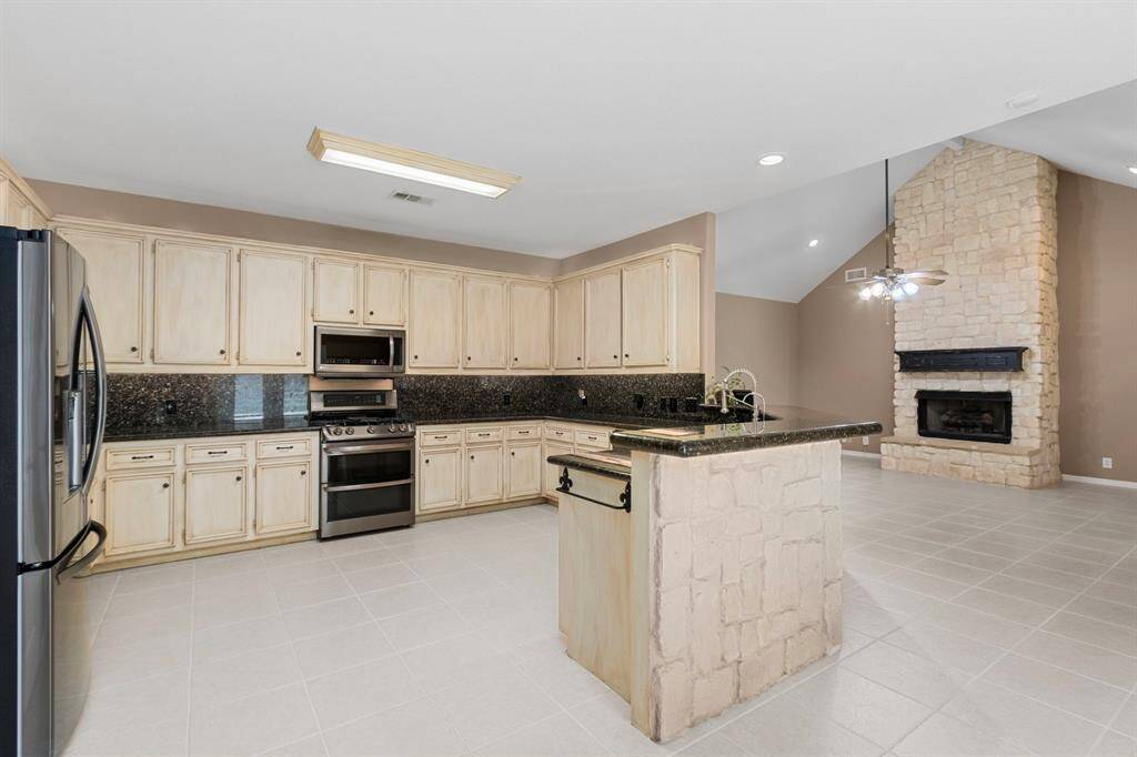 This spacious kitchen features light cabinetry, granite countertops, and stainless steel appliances. It opens to a living area with a stone fireplace and high ceilings, creating a warm and inviting atmosphere.