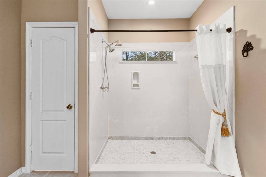 Spacious walk-in shower with a light neutral color scheme, featuring a high window for natural light and a curtain for privacy. Adjacent is a white door with classic detailing.