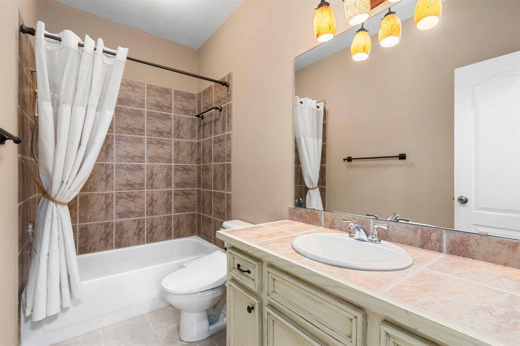 Bright bathroom with neutral tones featuring a tiled bathtub and shower combination. It includes a large mirror above a vanity with a tiled countertop and a modern sink. Warm lighting fixtures add a cozy touch.