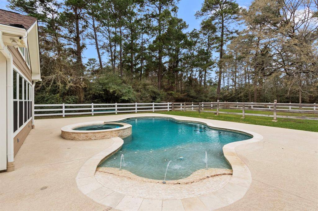 backyard with a uniquely shaped pool featuring a built-in hot tub. The area is surrounded by a fence and bordered by lush trees, providing a private and serene atmosphere. The patio is spacious, ideal for outdoor relaxation and entertainment.