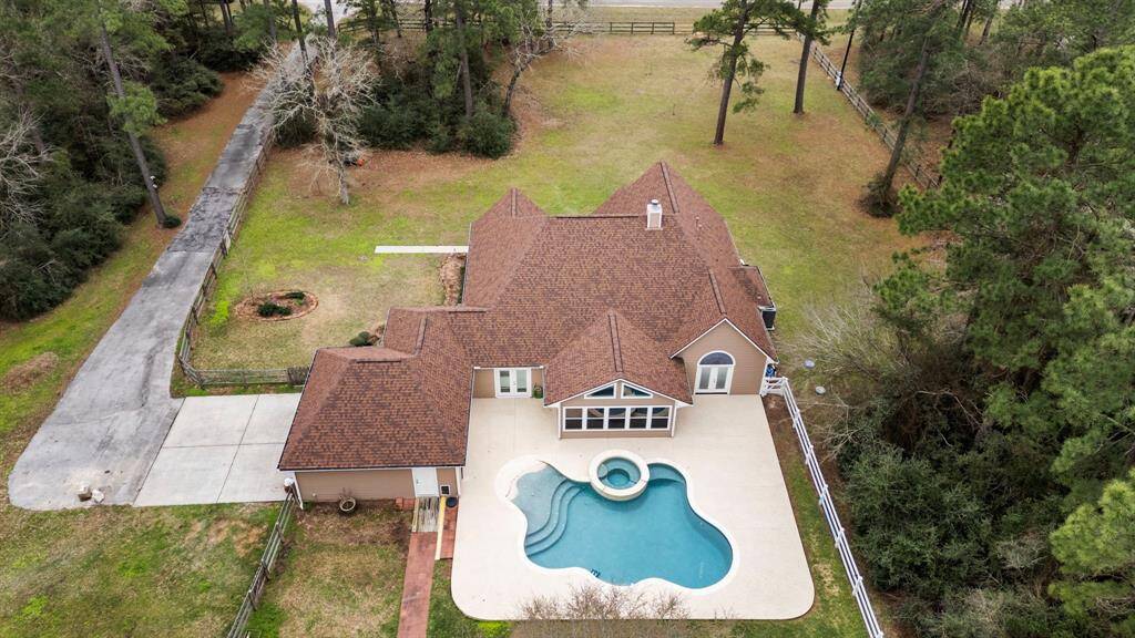 This property features a spacious single-story home with a brown roof, a large driveway, and a landscaped yard. It boasts a backyard pool with an attached spa, surrounded by a patio area, and is nestled in a serene, wooded setting for added privacy.