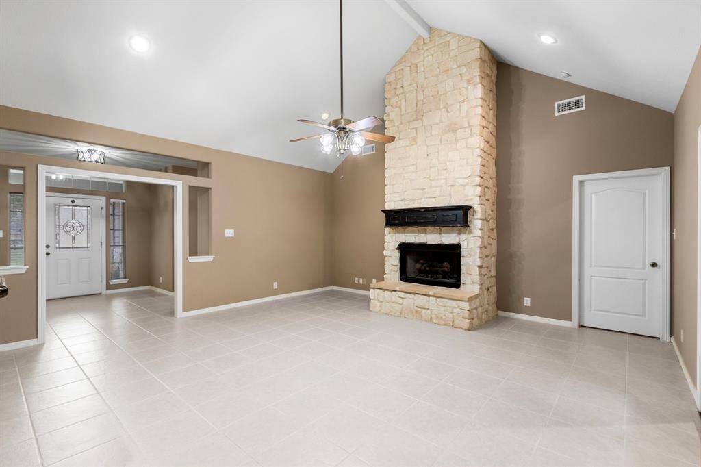 This spacious living area features a vaulted ceiling, neutral tile flooring, and a striking stone fireplace. The open layout flows into the entryway, with ample natural light from large windows and a decorative glass front door.