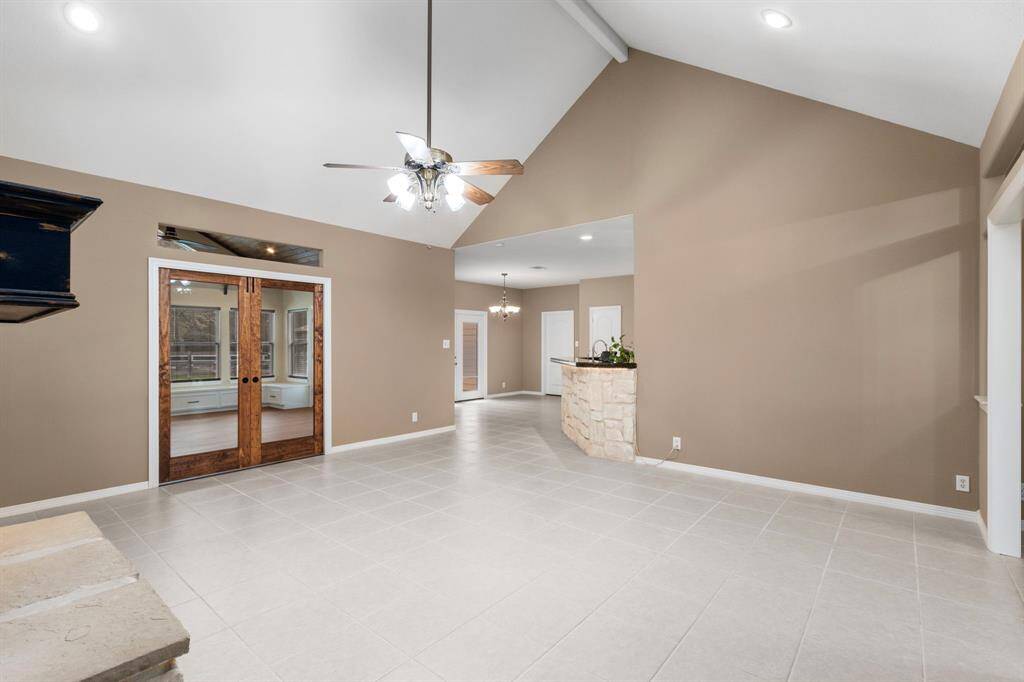 This spacious living area features high ceilings with a ceiling fan, neutral-toned walls, and a tile floor. The room is well-lit, with French doors leading to an adjacent space, and a stone-accented eat at bar counter space that adds a touch of character.