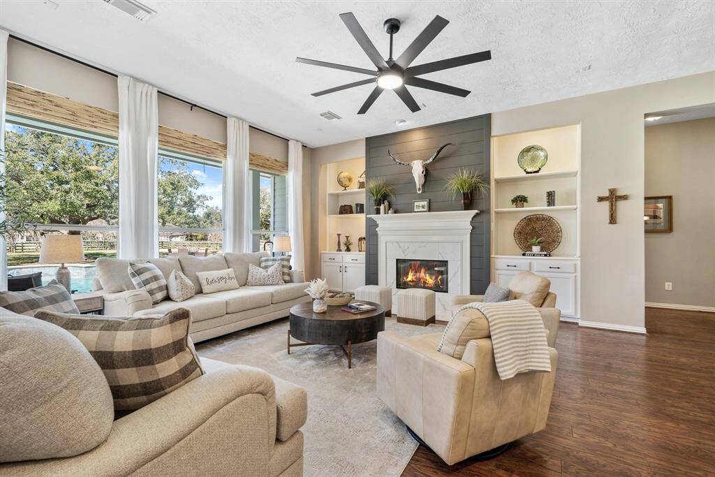 The Living Room boasts the high-end laminate flooring, a quartet of windows with bamboo-woven blinds, and a high-end, 8-blade ceiling fan. The hallway seen to the right leads to the Primary Retreat and Secondary Bedroom wing.