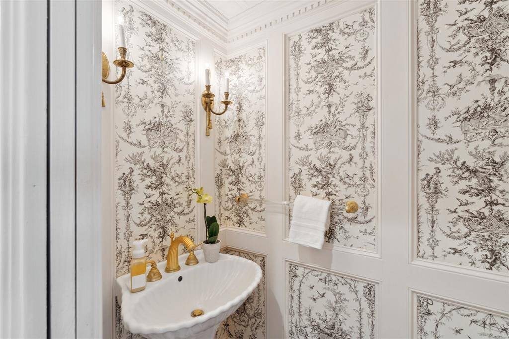 The charming powder bathroom [5' x 3'] is situated on the first floor across from the staircase and between the living and dining areas. The bathroom is nicely appointed by delicate dentil molding and beveled wood paneling. The walls are lined with toile wallpaper accented by decorative gold sconces. Lovely!