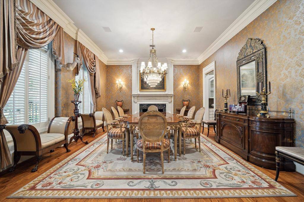 Dining room with oak hardwood floors with mahogany wood inlay, brass and crystal chandelier, dual brass sconces flanking the fireplace, recessed lighting, custom millwork, wood-burning fireplace with a custom wood mantle, marble surround and gas logs, plantation shutters, custom drapery, and built-in speakers.