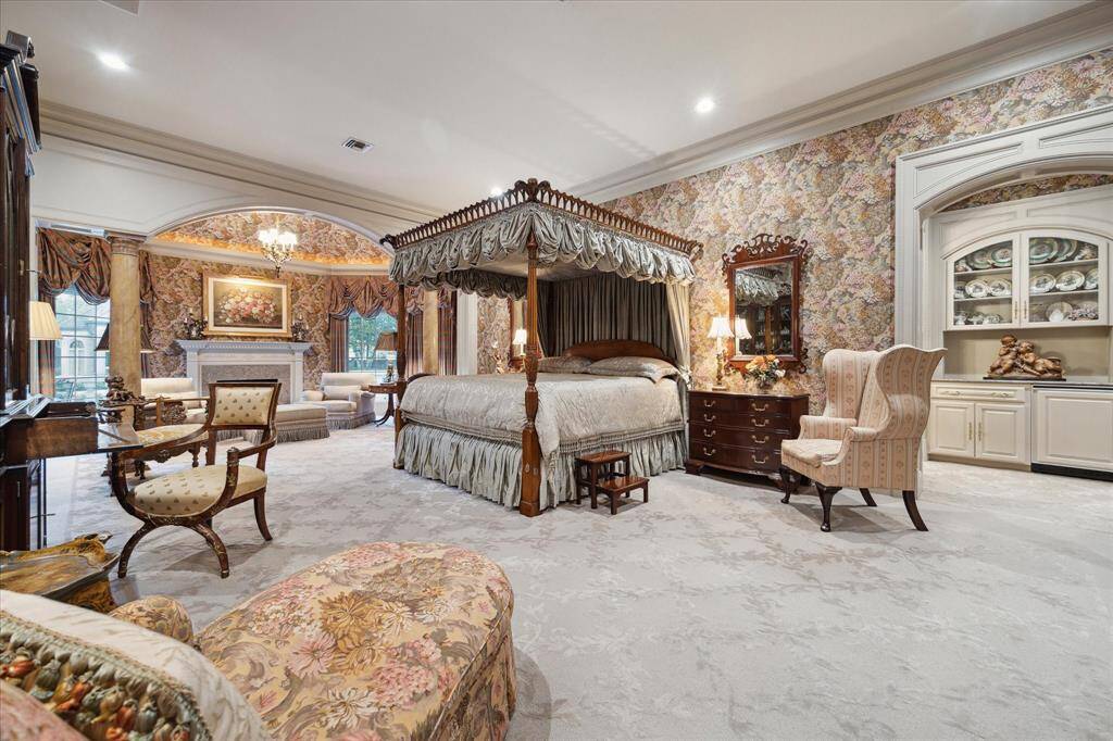 Primary suite with Hokanson carpeted flooring, an ornate brass chandelier, recessed lighting, faux-finished columns leading to the sitting area, custom drapery, octagonal vaulted ceiling, wood-burning fireplace a custom wood mantle and marble surround accented by andirons, views to the rear grounds, door to the outside, built-in coffee bar with a refrigerator, pocket doors to the separate dual bathrooms, and built-in speakers.