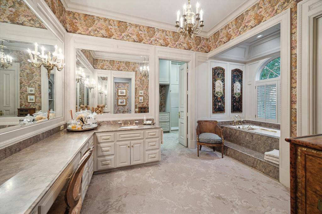 First primary closet with carpeted flooring, florescent lighting, oculus window, custom millwork, built-in  drawers and shelving, marble countertops, brass hardware, abundant hanging space, and valet rods.