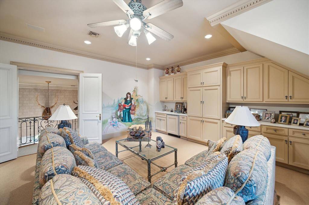 Family room with carpeted flooring, recessed lighting, ceiling fan with light, custom millwork, plantation shutters, custom drapery, window seating with built-in storage, built-in cabinetry with pewter hardware, wet bar with solid surface counters, ceramic sink, brass plumbing fixtures, built-in beverage refrigerator, and built-in speakers.