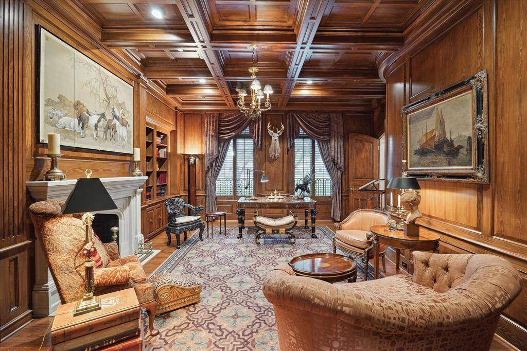 Library with oak hardwood floors, ornate brass chandelier, recessed lighting, paneled boxed beam ceiling, custom oak paneling and millwork, wood-burning fireplace with a limestone mantle and surround accented by brass and iron andirons, plantation shutters, custom drapery and built-in speakers.