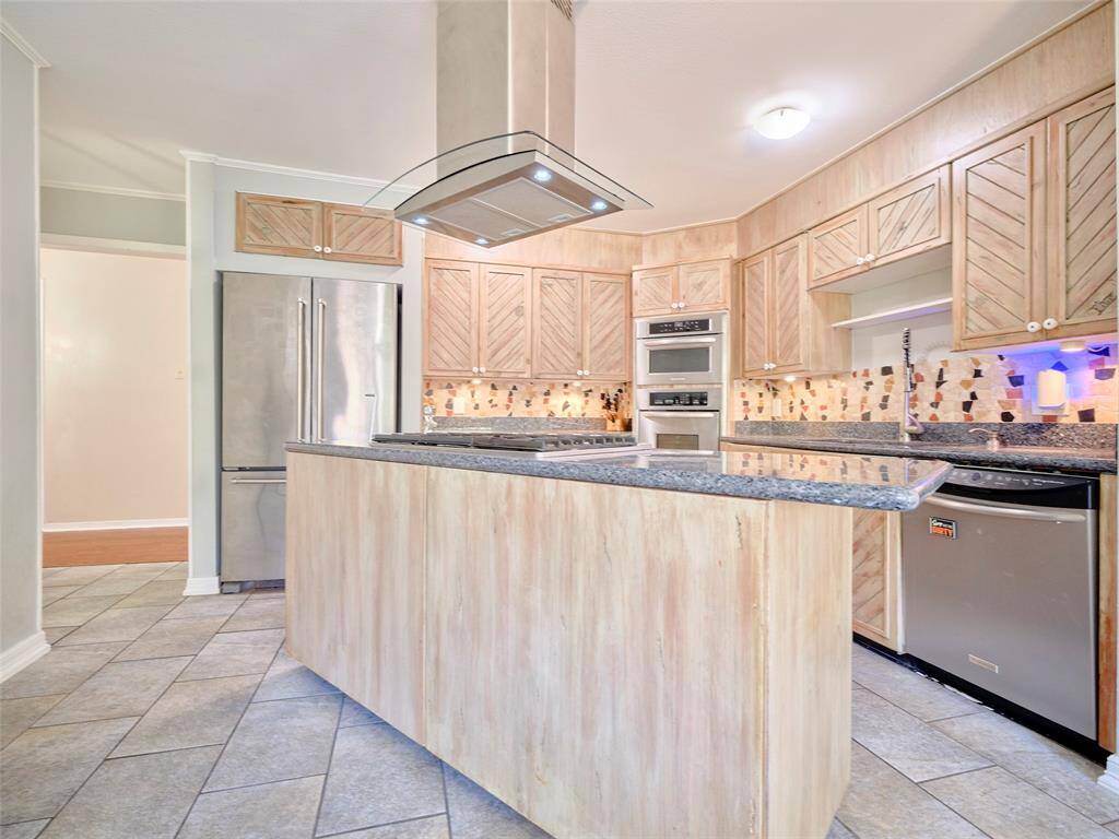 This kitchen features light wood cabinetry, stainless steel appliances, a central island with a cooktop, and a stylish backsplash. The floor is tiled, and the space is well-lit with natural and artificial lighting.