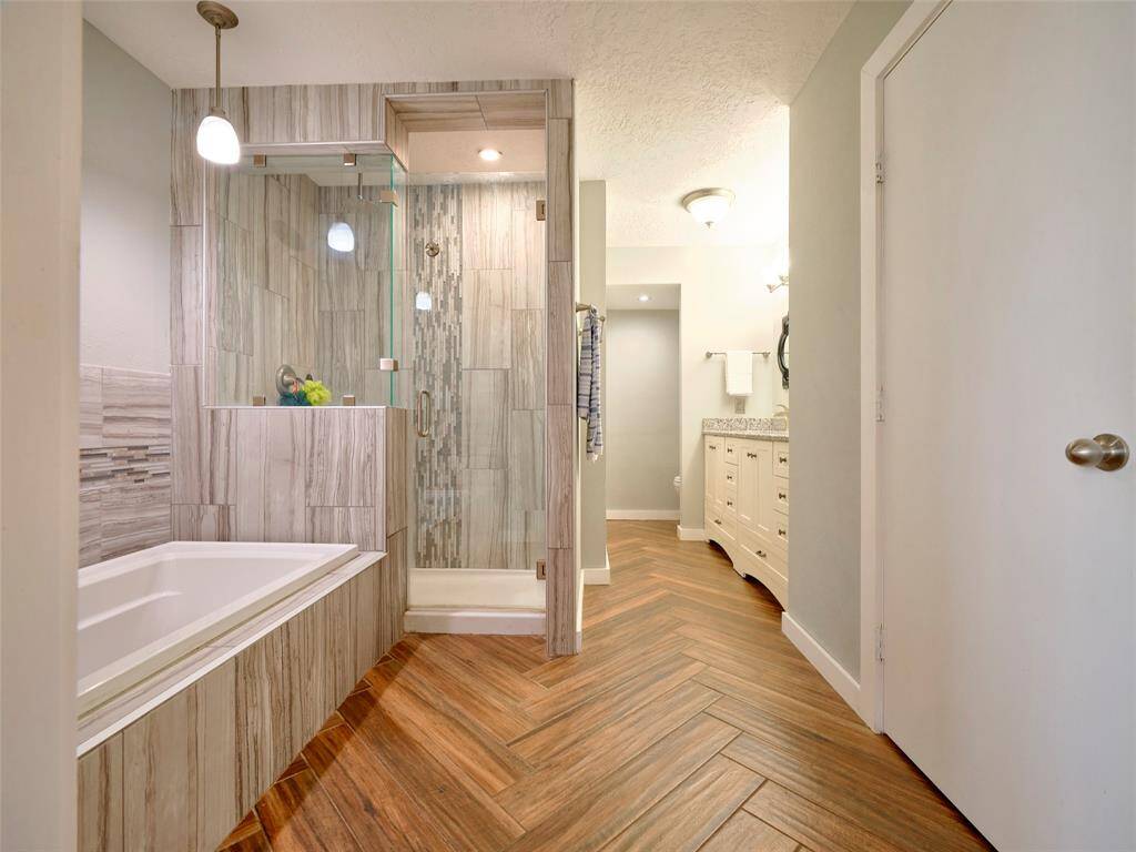 This bathroom features modern wood-look tile flooring, a large walk-in shower with glass doors, and a separate bathtub. The vanity area has ample storage and is well-lit.