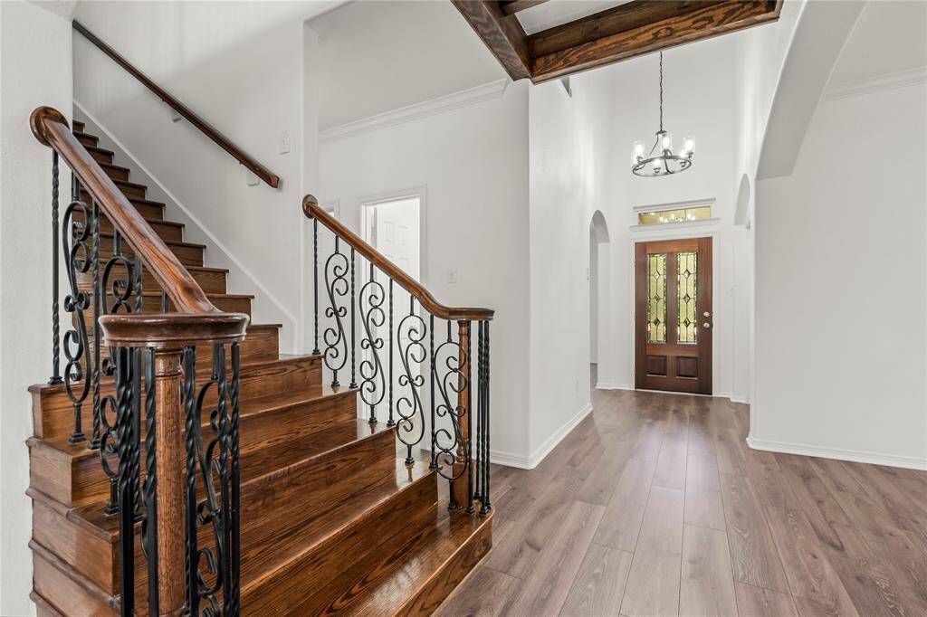This inviting entryway features elegant plank vinyl flooring and a grand staircase with ornate iron railings. The space is bright and airy, highlighted by a stylish chandelier and a beautiful wooden front door with decorative glass panels.