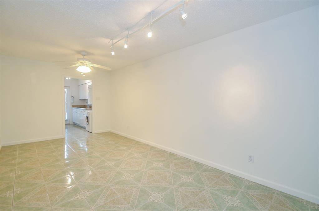 This bright room features tiled flooring with a geometric pattern, a ceiling fan, and track lighting. It leads into a kitchen area with visible appliances and white cabinetry, creating an open and inviting space.