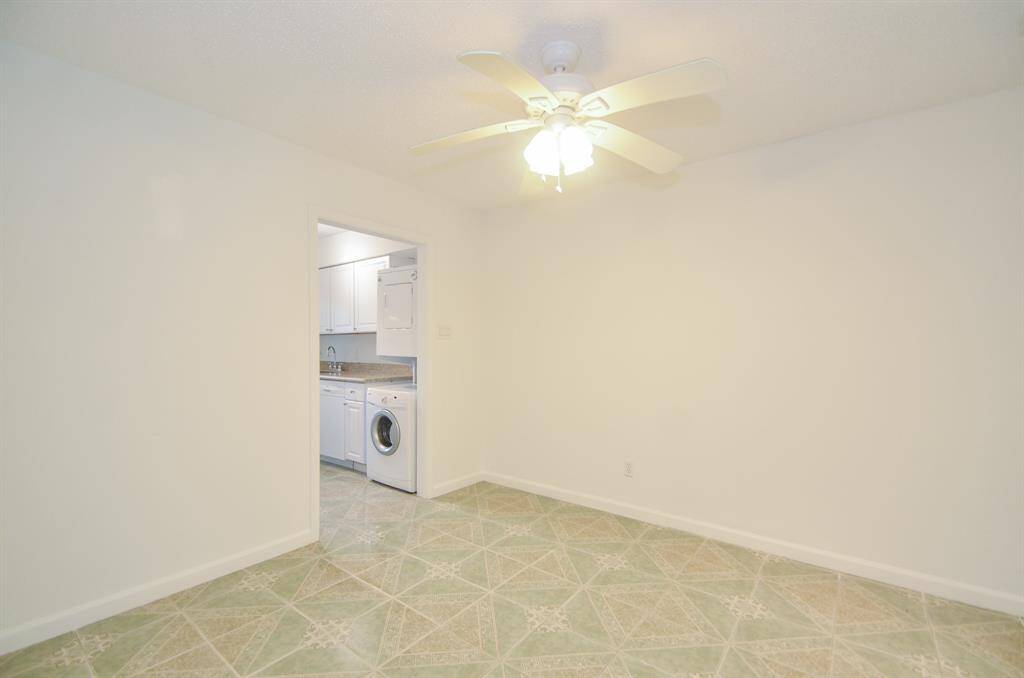 Bright room with tiled flooring and a ceiling fan. Adjacent to a kitchen area featuring white cabinets, a sink, and a stacked washer-dryer unit.