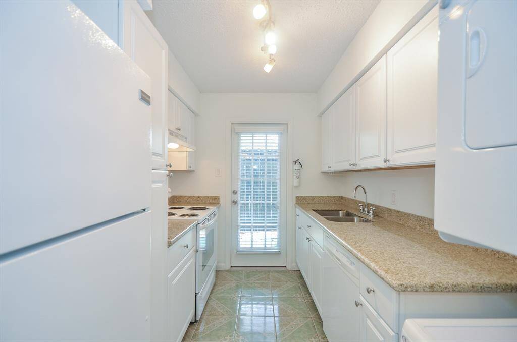 This kitchen is compact and bright, featuring white cabinetry and appliances, granite countertops, and a tiled floor. A glass door at the end brings in natural light.