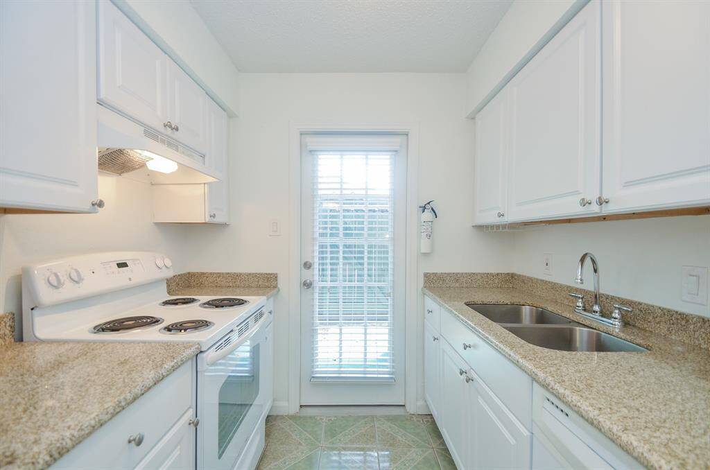 This photo shows a compact, bright kitchen with white cabinetry and granite countertops. It features an electric stove, dual sink, and a door with glass panels leading outside, enhancing natural light.