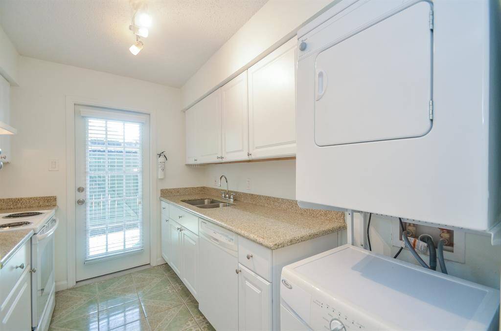 This bright and compact utility room features white cabinetry, granite countertops, and a stackable washer and dryer. A door with glass panels leads to the outside, providing natural light and easy access.