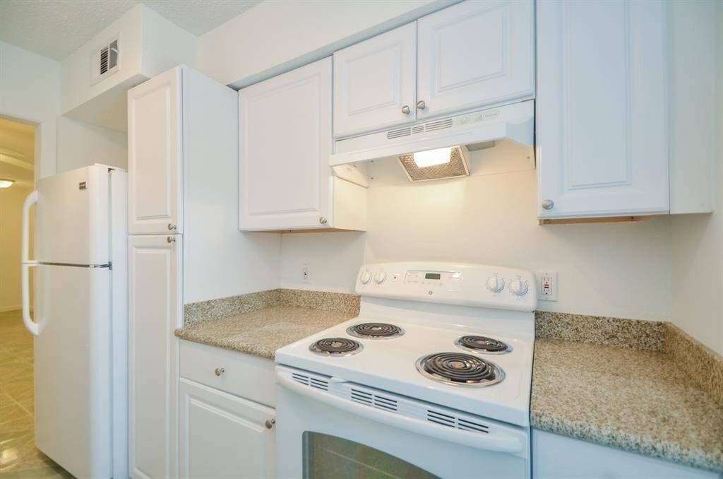 This kitchen features white cabinetry, a white electric stove with a range hood, granite countertops, and a refrigerator, offering a clean and functional cooking space.