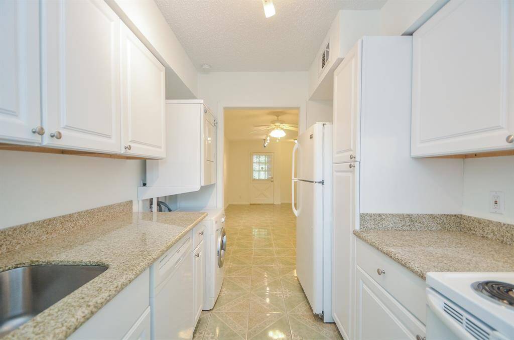 This photo shows a bright, narrow kitchen with white cabinets and granite countertops. It features a sink, appliances, and a washer/dryer. The kitchen leads into a room with a ceiling fan and natural light.