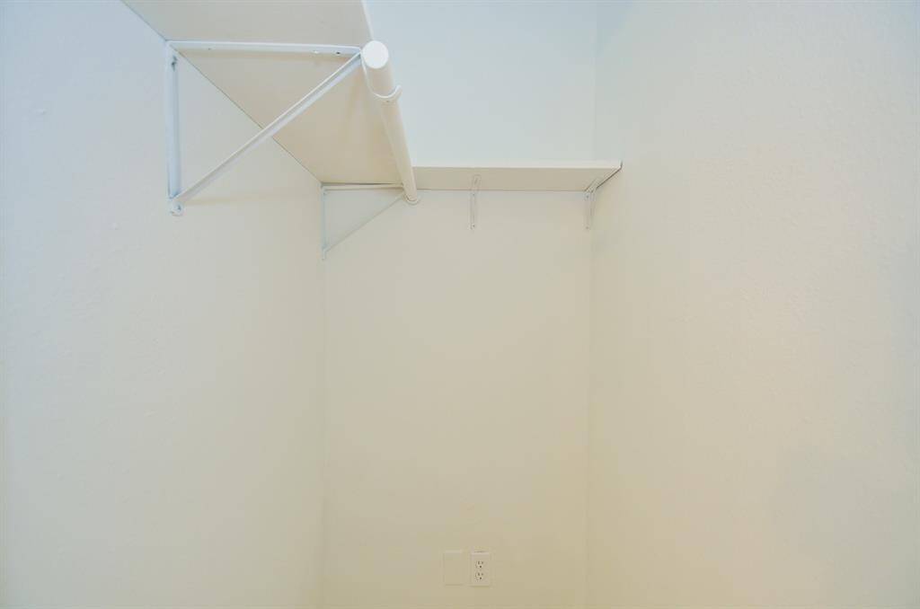 This photo shows a small, white closet space with a single shelf and a hanging rod, ideal for additional storage or organizing clothing. An electrical outlet is visible on the wall.