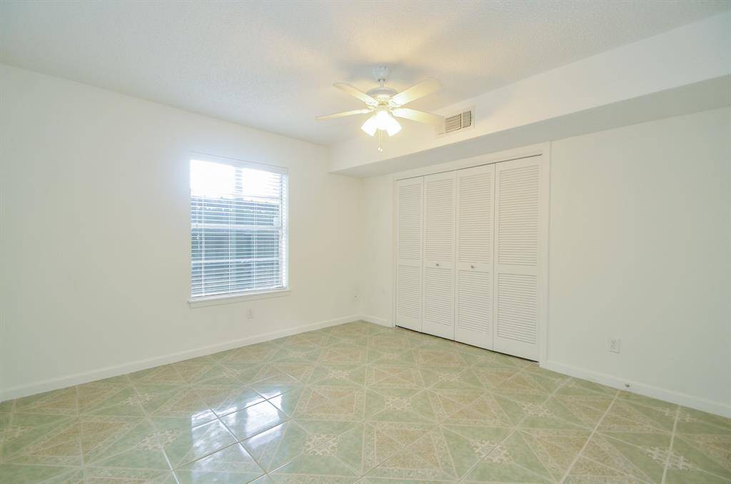 A bright, spacious room featuring tiled floors, a ceiling fan, and a large window with blinds. It includes a white closet with double doors, offering ample storage.