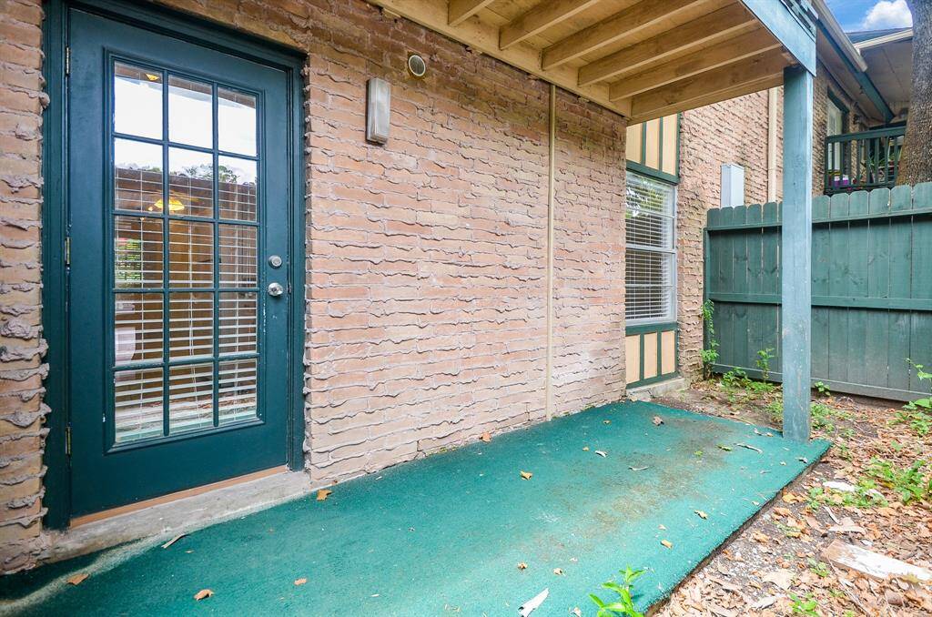 This photo shows a cozy outdoor patio area with a green carpet, enclosed by a brick wall and wooden fencing. It features a glass-paneled door leading inside, ideal for a small seating area or garden space.