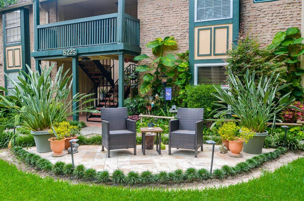 Charming outdoor patio area with two wicker chairs and a small table, surrounded by lush greenery and potted plants. The setting offers a cozy and inviting space for relaxation. The exterior features brick siding and a small balcony, adding to the quaint appeal.