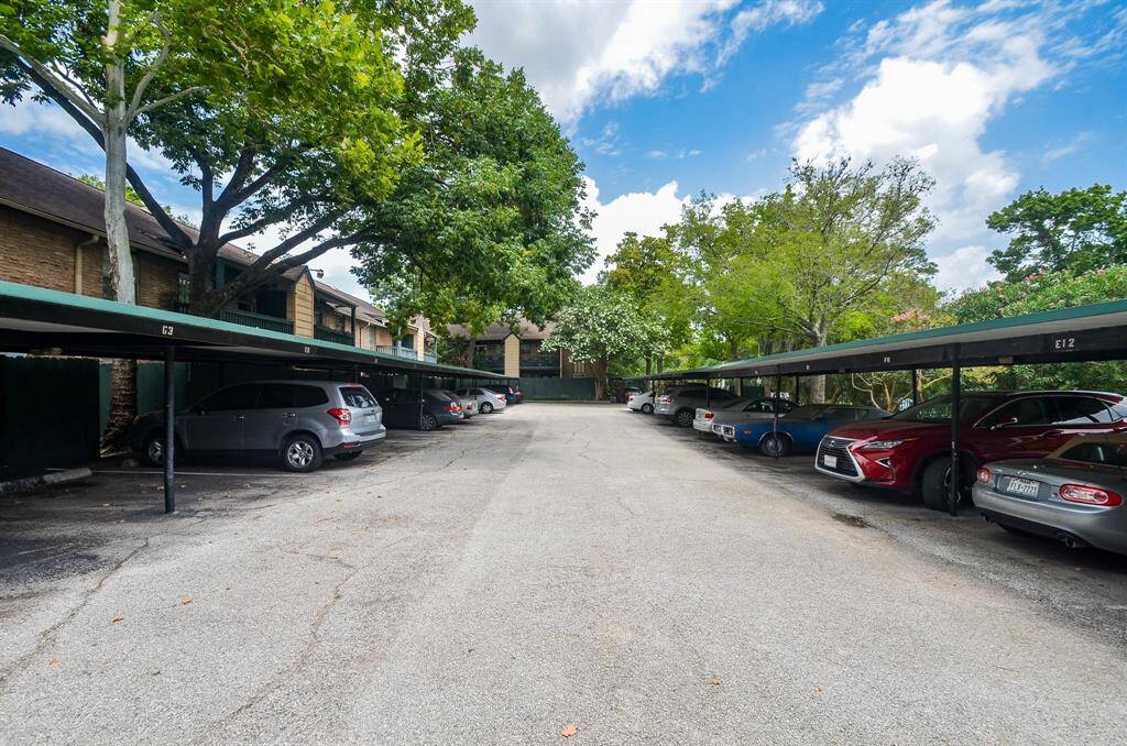 This photo shows a covered parking area with spaces for multiple cars, surrounded by trees and adjacent to a residential building. The setting indicates a quiet, well-maintained community.