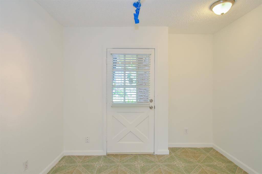 A small, well-lit room with white walls and a patterned tile floor. It features a door with a window, allowing natural light to enter, and a simple ceiling light fixture.