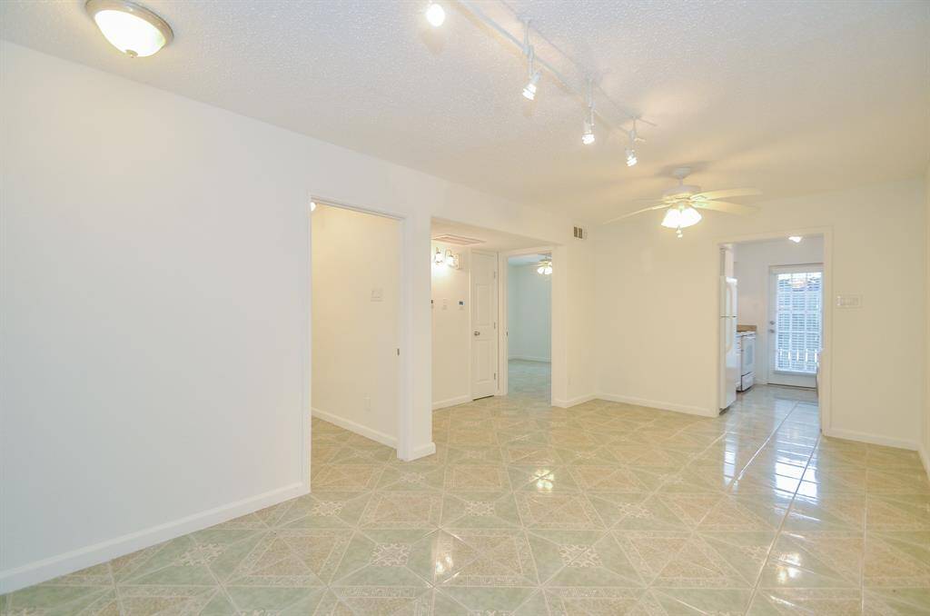 This photo shows a bright, open living area with tiled flooring and neutral walls. It features a ceiling fan, track lighting, and leads into a kitchen with visible appliances. The space offers a clean, versatile layout for various furniture arrangements.