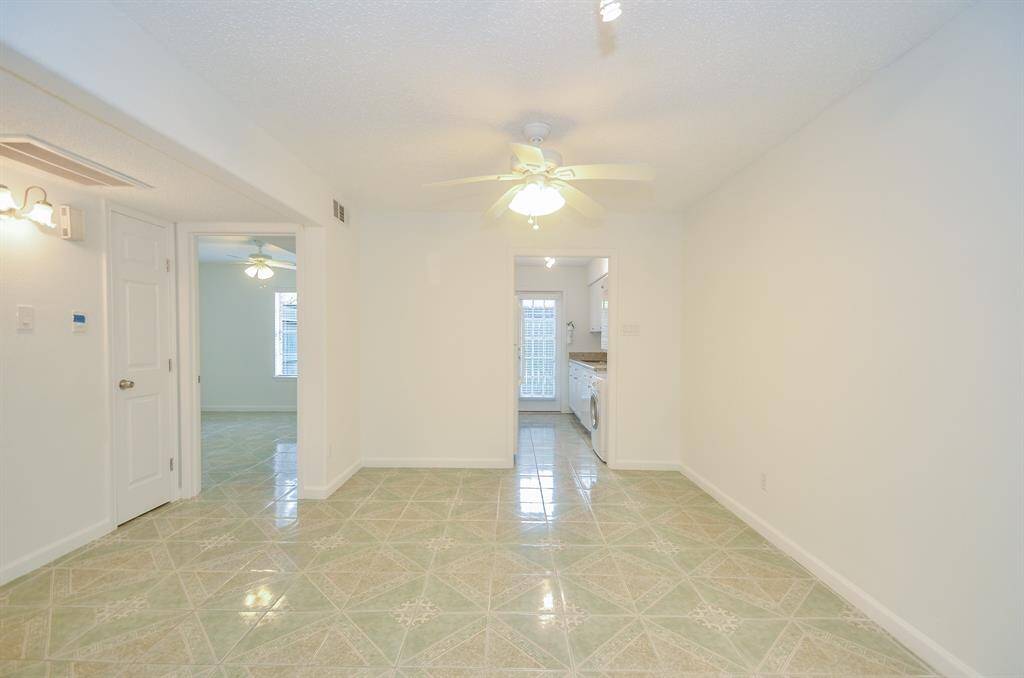 This photo shows a bright, clean interior with tiled flooring and a ceiling fan. The space leads into another room with a similar setup, and a kitchen area with a washer and dryer is visible in the background. White walls and ample lighting create an open and inviting atmosphere.