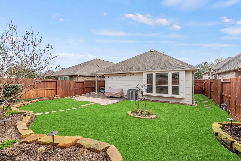 This inviting backyard offers a covered patio with a flagstone extension, creating the perfect space for outdoor gatherings. A lush lawn with sprinkler system is surrounded by rock-lined beds with mature shrubs and landscape lights providing soft ambient lighting in the evenings.