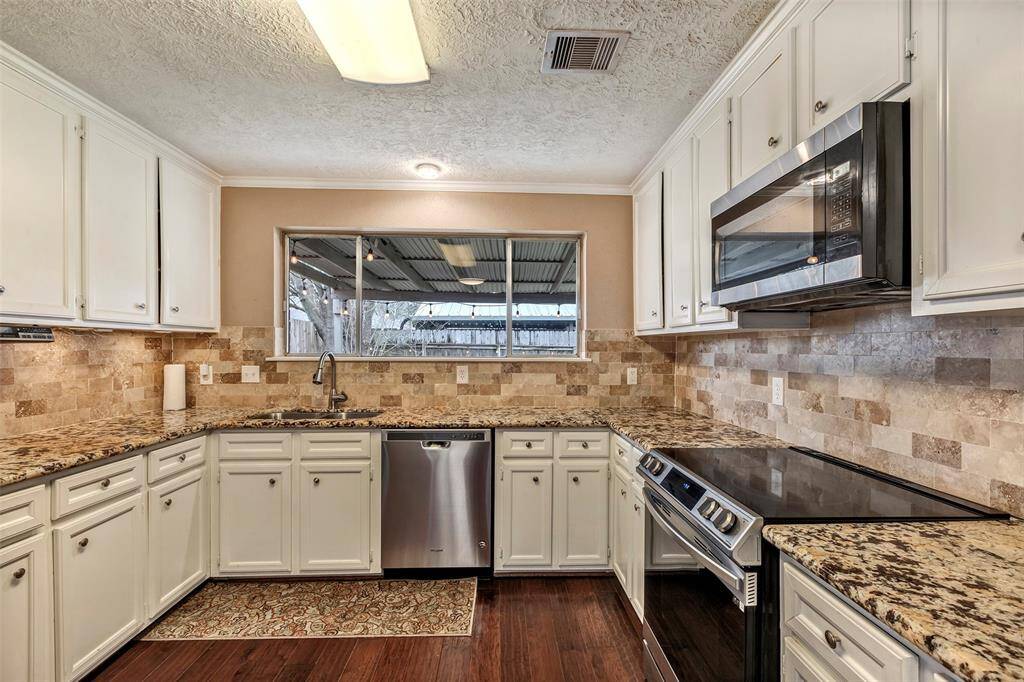 This kitchen features a modern design with granite countertops, white cabinetry, and stainless steel appliances. The backsplash is a stylish stone tile, and there's a large window for natural light. The dark wood flooring adds warmth to the space.