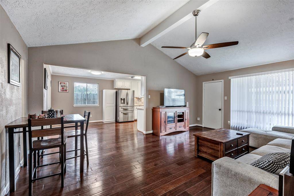 This photo showcases a bright, open-concept living and dining area with hardwood floors, vaulted ceilings, and a ceiling fan. The room features a cozy seating area, a dining table, and connects seamlessly to a modern kitchen with stainless steel appliances. Large windows and sliding doors provide ample natural light.