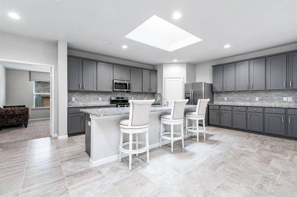 This spacious kitchen features modern gray cabinetry, a large central island with seating, and sleek stainless steel appliances. The room is brightened by a skylight and recessed lighting, with a neutral tile floor that complements the stylish design.