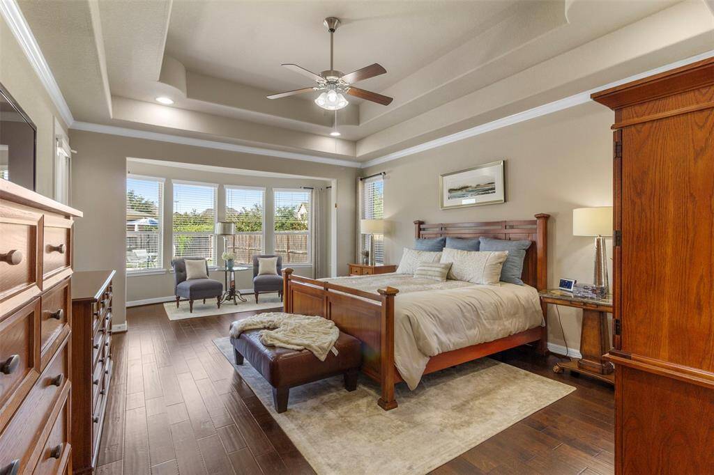 Step into the sanctuary of theprimary bedroom featuring wood floors, high tray ceilings, recessed lighting, ceiling fan, crown molding, sitting area, and large windows.