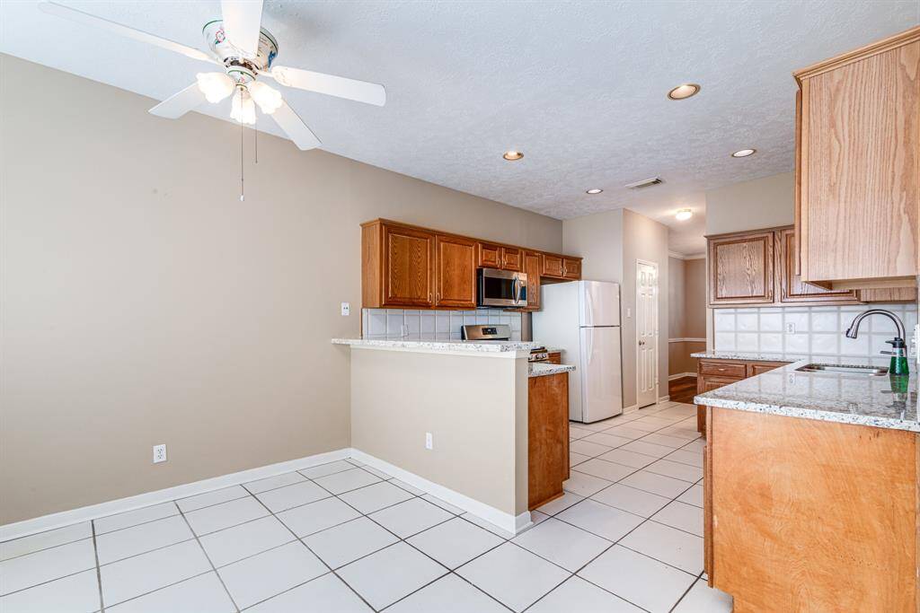 Bright kitchen with spacious layout, featuring white tile flooring and neutral walls. It has wooden cabinets, stainless steel appliances, and granite countertops. Ceiling fan adds comfort to the open dining area.