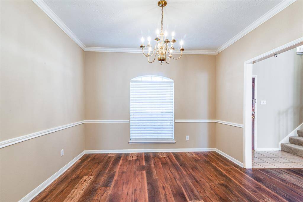 This photo shows a bright, inviting dining room with warm wood flooring, neutral walls, and elegant crown molding. A classic chandelier hangs from the ceiling, and a window with blinds allows natural light in.
