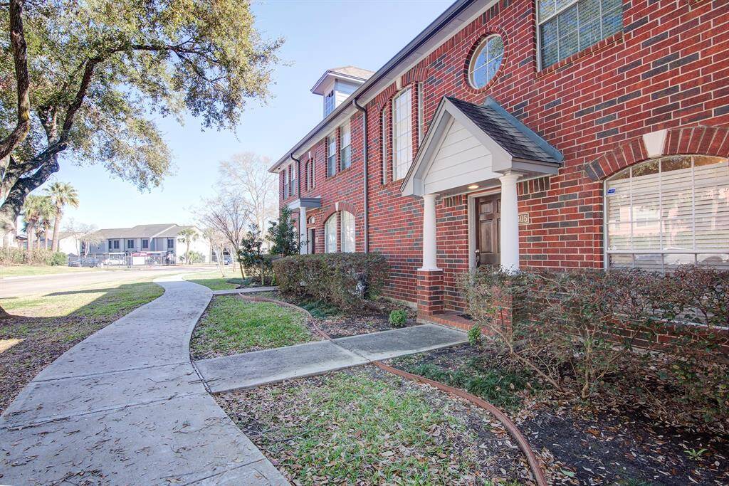 Brick townhome with a classic exterior, featuring a welcoming front entrance with white columns. A curved walkway leads through a landscaped area with mature trees. Ideal for those seeking a charming, well-maintained home in a pleasant neighborhood.