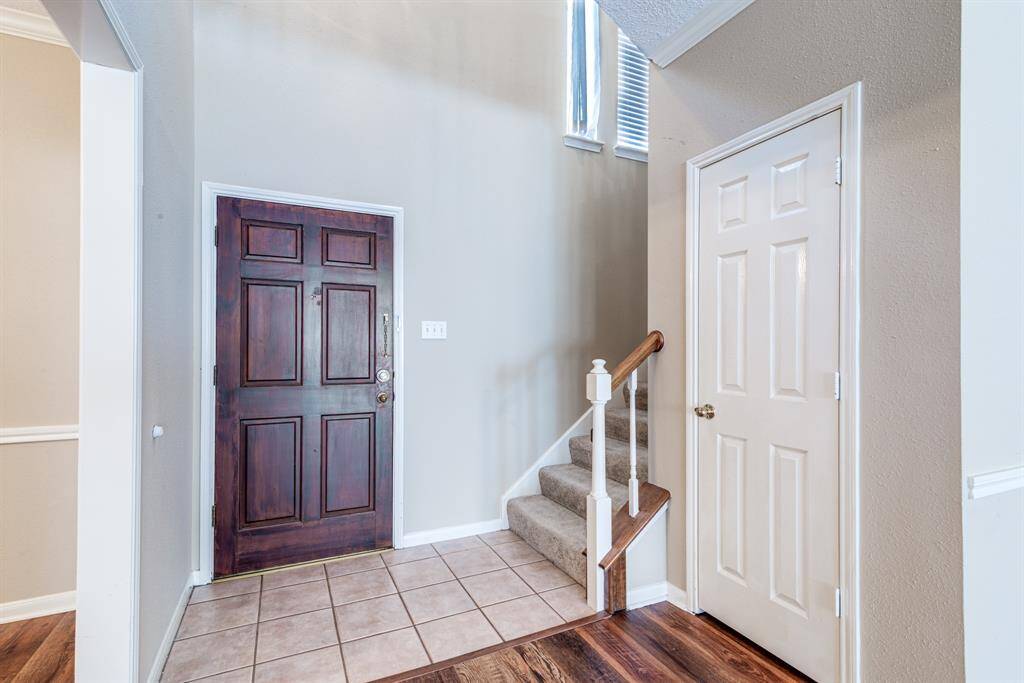 This entryway features a classic wooden front door, tile flooring, and a staircase with carpeted steps. The neutral wall color is complemented by natural light, creating a warm and inviting space.