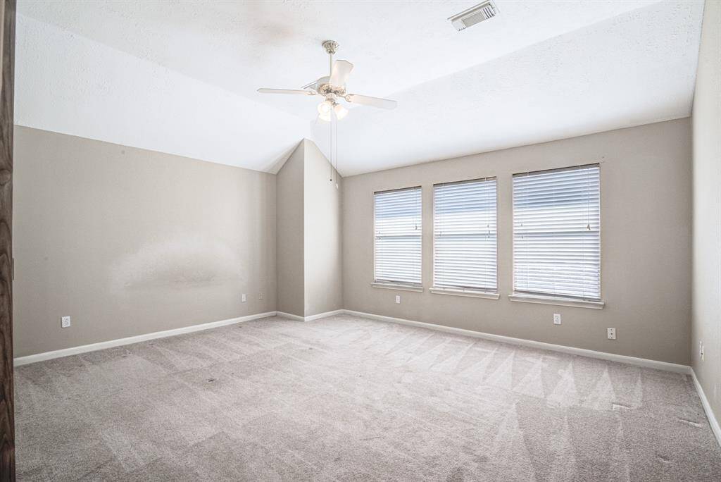Spacious primary bedroom with neutral walls and carpet, featuring a vaulted ceiling and a ceiling fan. Large windows provide ample natural light, enhancing the room's airy feel.