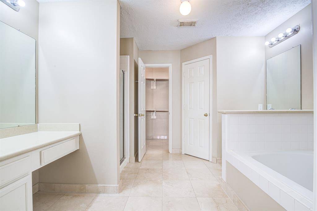 This spacious primary bathroom features a large vanity on your left and a second vanity on the right with ample counter space, a soaking bathtub, and a separate shower. Neutral tones and bright lighting create a clean, inviting atmosphere, complemented by a walk-in closet for added convenience.