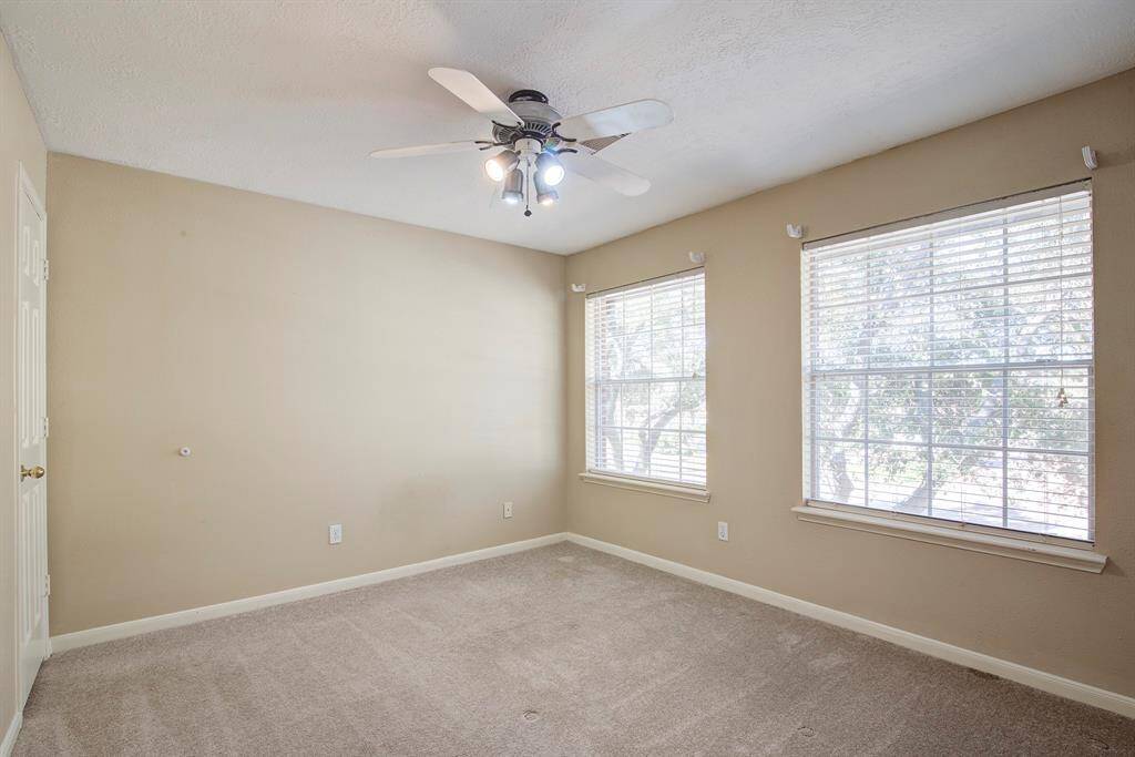 This bedroom features neutral walls, a ceiling fan with lights, and two large windows that provide ample natural light. The space is carpeted, offering a cozy feel, and is ideal for a bedroom or home office.