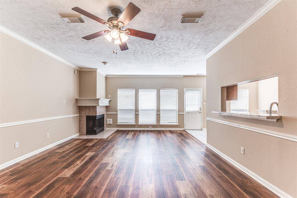 This spacious living area features hardwood flooring, a cozy corner fireplace, and abundant natural light from large windows. The open layout includes a ceiling fan and a convenient pass-through to the kitchen, ideal for entertaining.