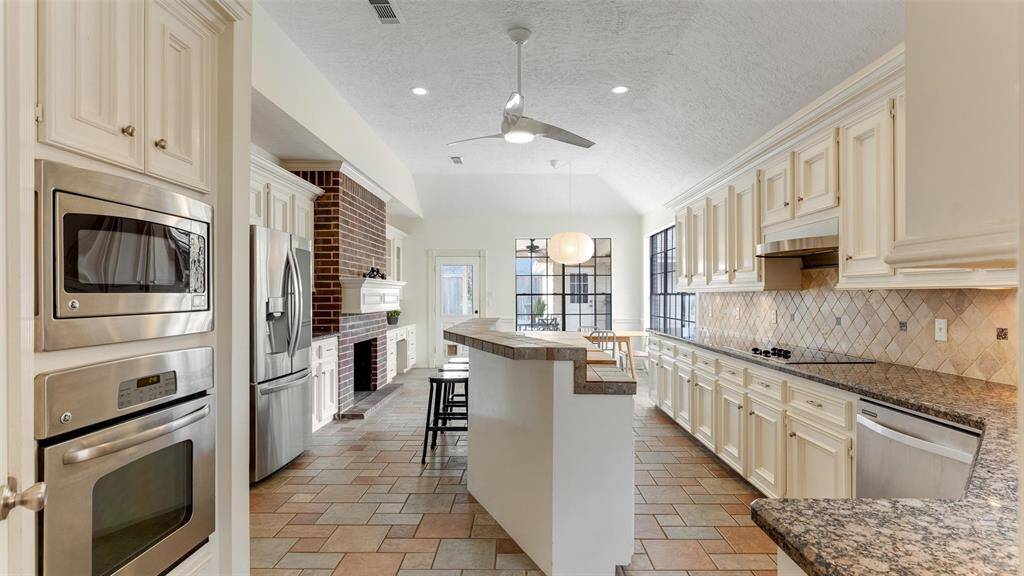 This expansive kitchen boasts a long granite countertop, abundant cabinetry, and sleek stainless steel appliances. It includes a breakfast bar with seating, tiled flooring, and a cozy adjacent dining area with large windows that fill the space with natural light. A brick accent wall with a fireplace adds so much character.
